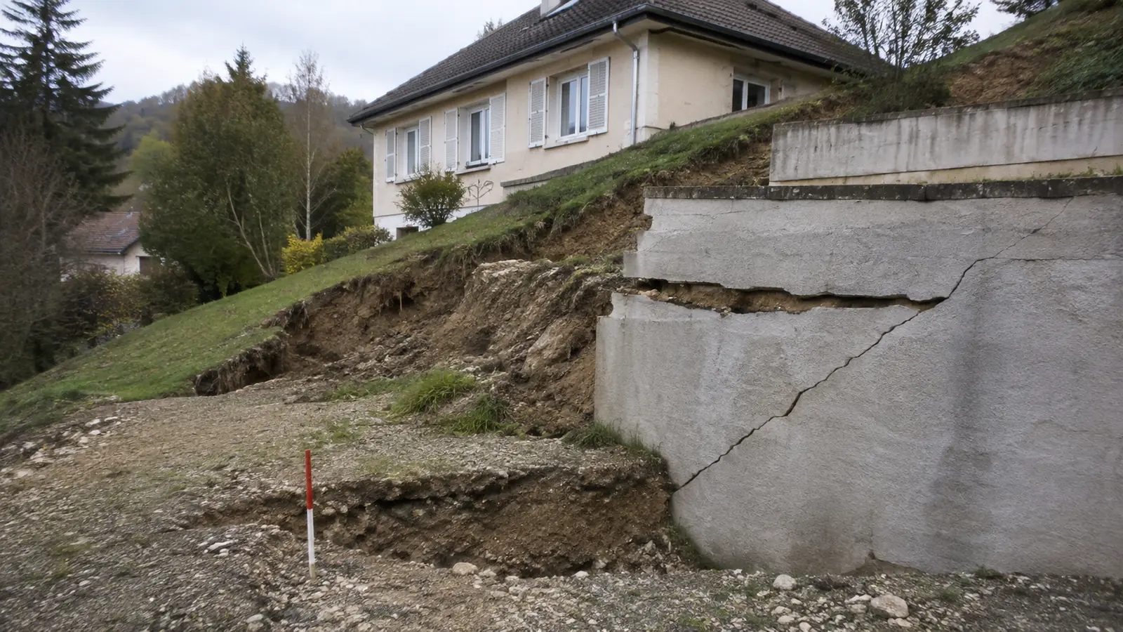 Mur de soutènement fissuré au pied d'un terrain en pente, sol affaissé et jalon de mesure planté au sol, maison intacte en arrière-plan